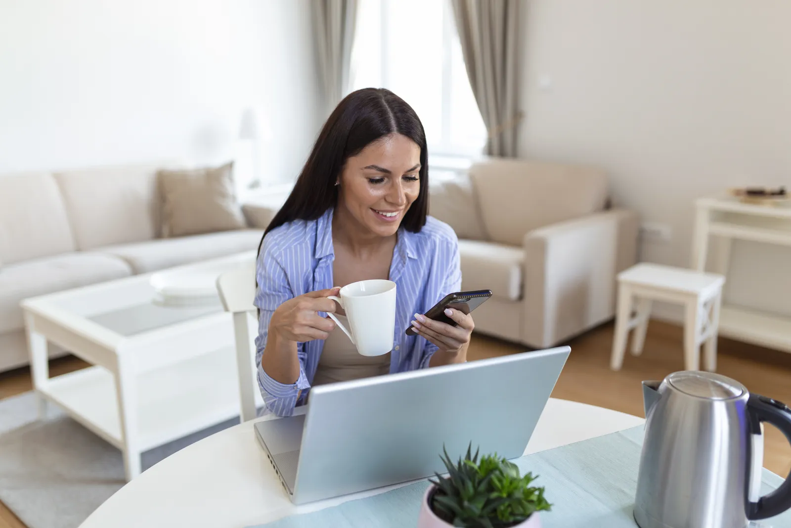 Donna al tavolo di casa con notebook aperto e smartphone in mano, accanto una tazza di caffè