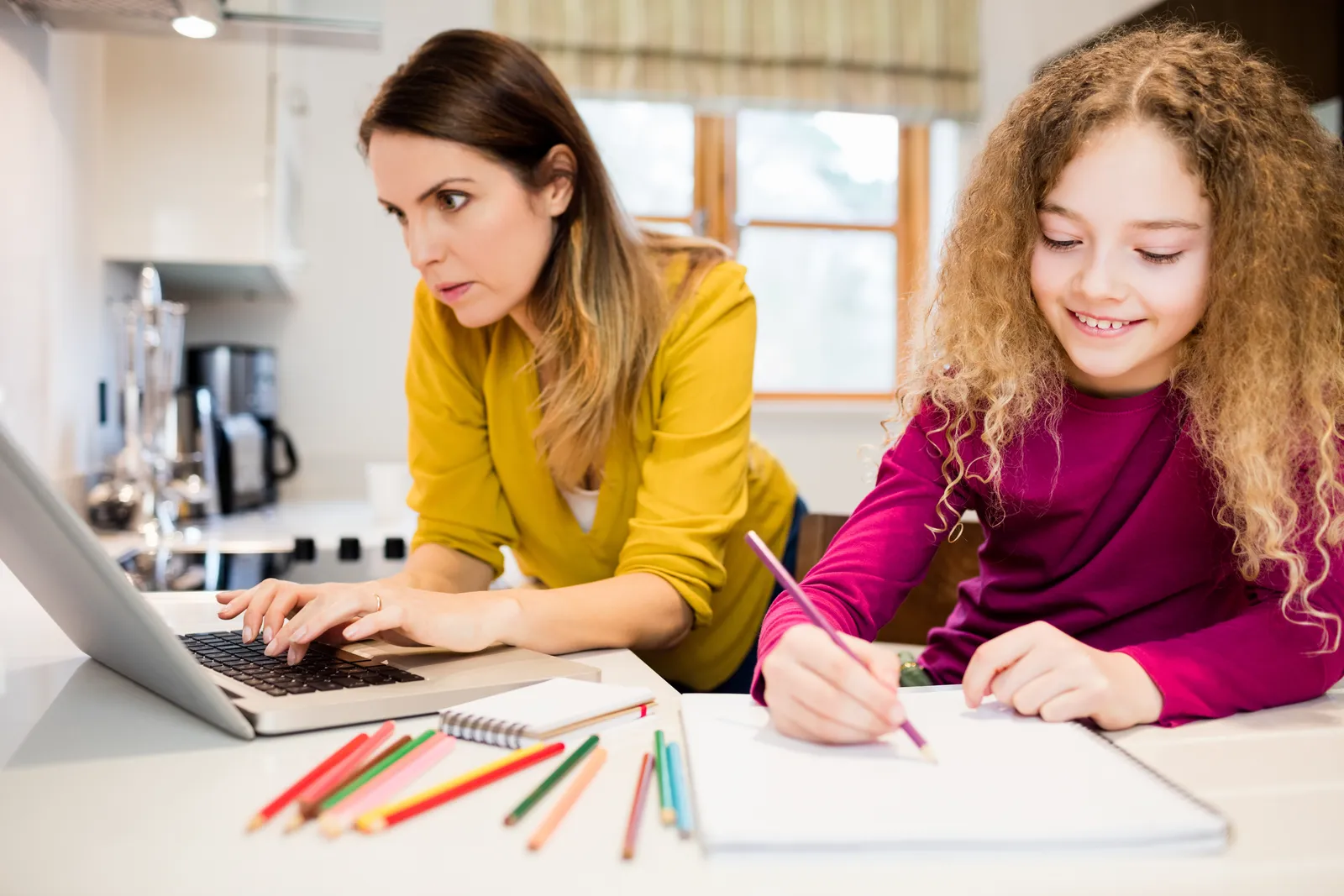 Madre che lavora al portatile in cucina mentre la figlia fa i compiti accanto a lei