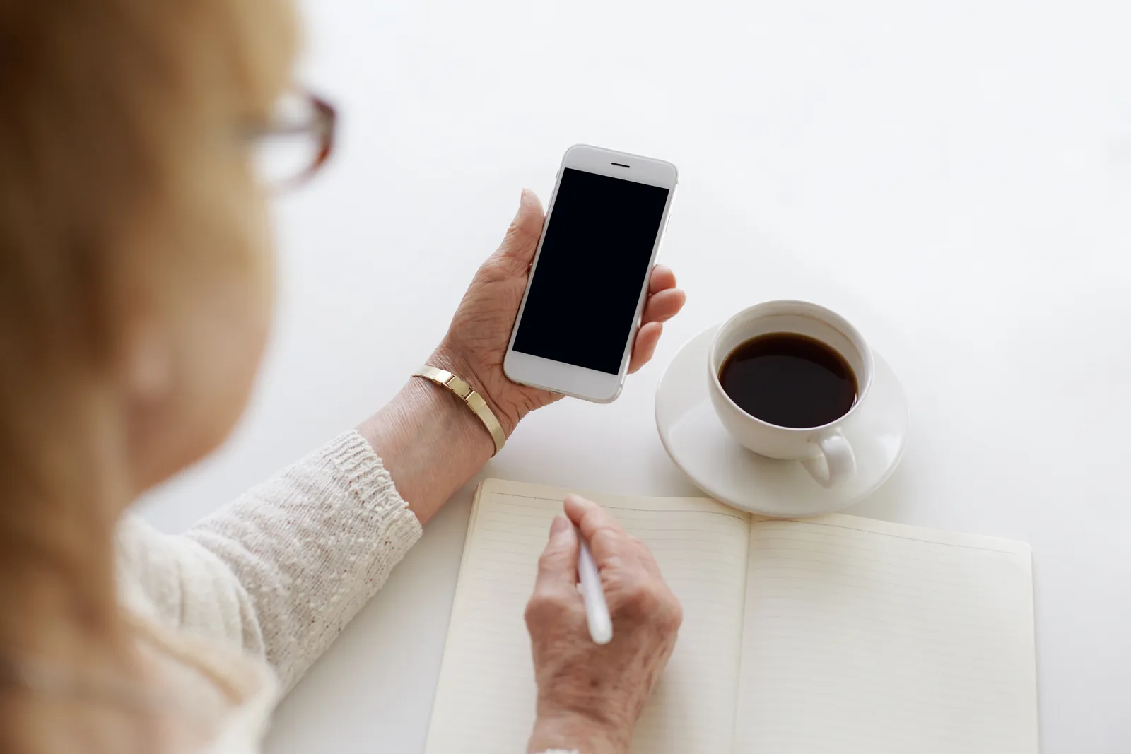 Mano che tiene uno smartphone sopra un tavolo con una tazza di caffè e un quaderno aperto